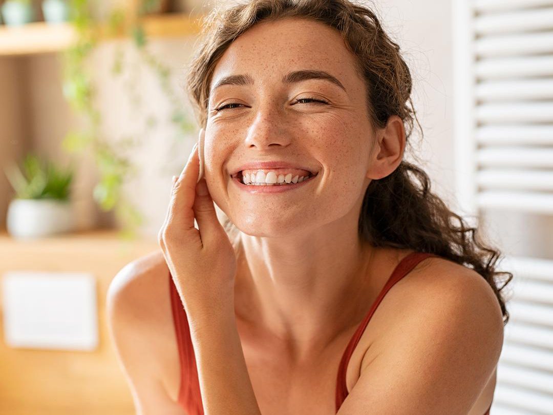 A woman with curly hair is smiling brightly and touching her face with one hand. She is wearing a sleeveless orange top. The background features a home setting with houseplants and wooden shelves, suggesting a relaxed and happy environment, perfect for those wondering, "What is sensitive skin?
