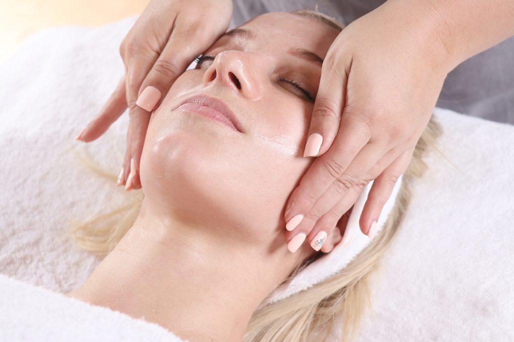 A person is lying down with a towel underneath their head, receiving a facial massage. The masseuse's hands are gently massaging the sides of the person's face with a relaxing touch.