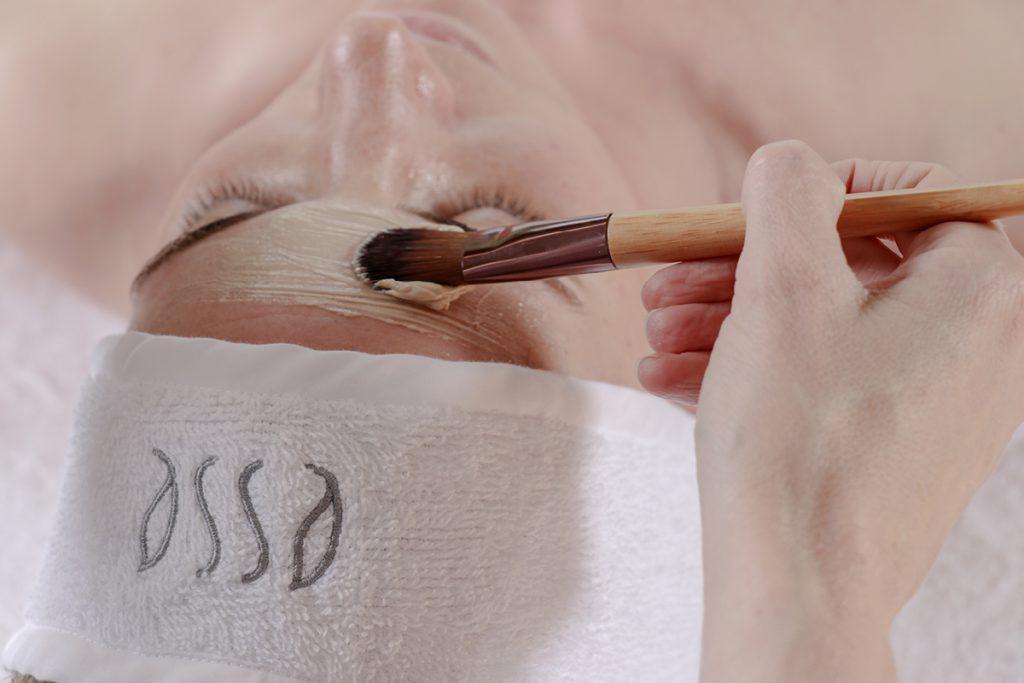 A person is lying down with a towel wrapped around their head. A close-up shows a hand holding a brush, applying a white creamy substance to the person's forehead, suggesting a facial treatment. The towel has the brand name "Esse" embroidered on it.