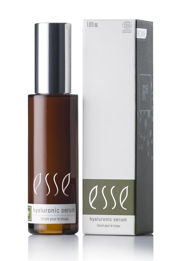 An Esse Hyaluronic Serum, encased in a brown bottle with a silver cap and labeled with white text, is displayed alongside its packaging. The box is predominantly white with one dark gray side, showcasing the same white text as on the bottle plus some extra details.