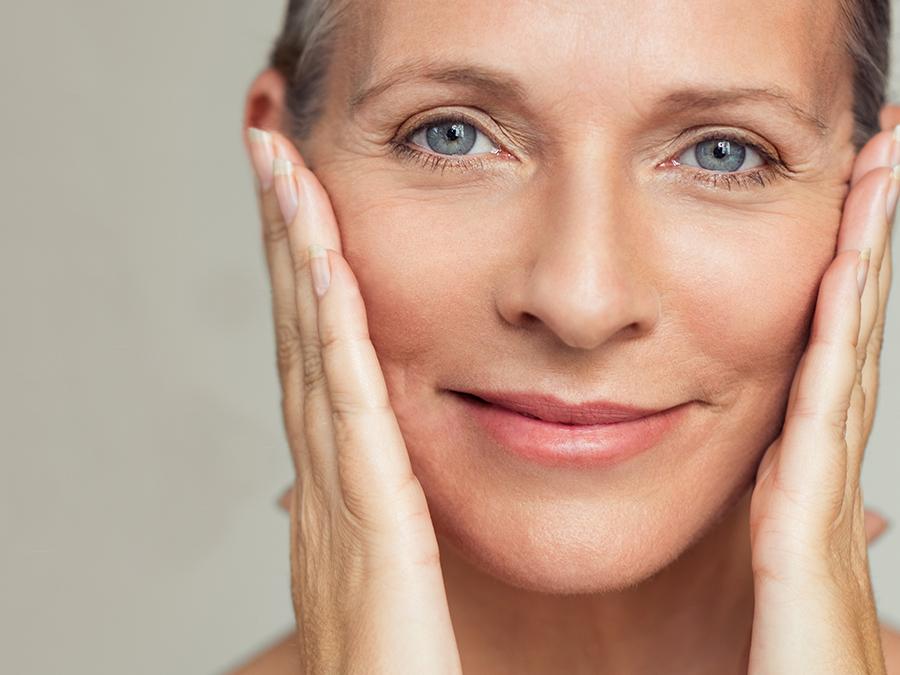 Close-up of a person with short, silver hair holding their face with both hands. They are smiling slightly, showcasing smooth skin and bright blue eyes. The background is a soft, neutral shade that complements the subject's serene expression.