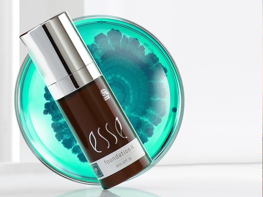 A sleek brown bottle with a silver pump dispenser labeled "concealer" is positioned in front of a turquoise, circular, artistic background. The bottle text includes "Esse" and "with SPF 30.