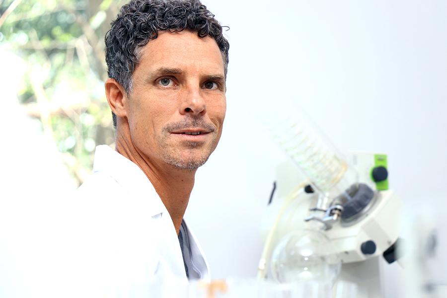 A person with short curly hair and a white lab coat stands next to scientific equipment, possibly in a laboratory setting. The background is bright and features an indistinct view of greenery outside the lab.