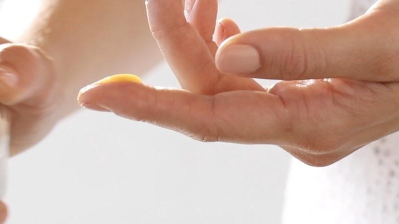 Close-up of a person's hands as they apply a small amount of Esse Ageless Serum to their fingertip. The image shows the serum being dispensed from a bottle, with the hands in focus against a blurred background.