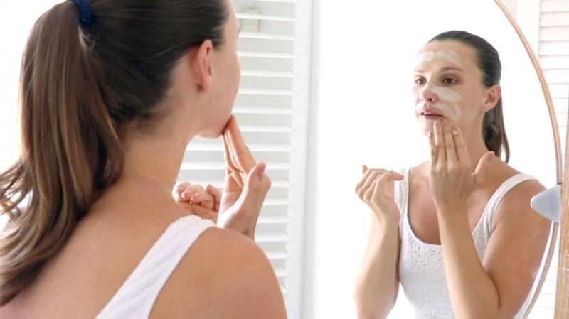 A person with long hair in a ponytail is applying the Esse Clay Mask while looking in the mirror. They are wearing a white tank top and are standing in a brightly lit bathroom with white shutters in the background.