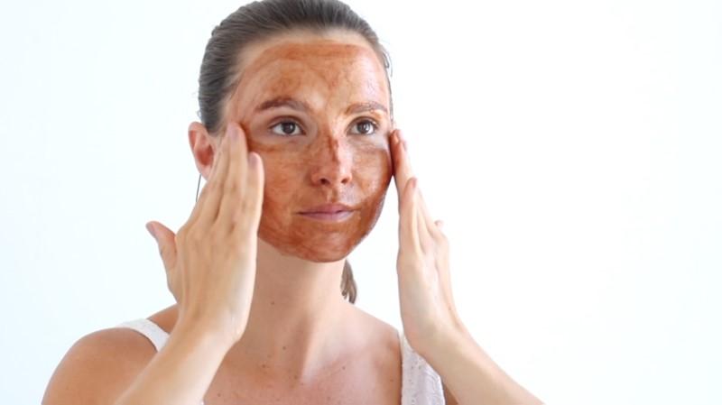 A woman is applying the Esse Cocoa Exfoliator to her face with both hands. She has brown hair pulled back and is wearing a white tank top. The background is white and minimalist, creating a clean and simple setting.