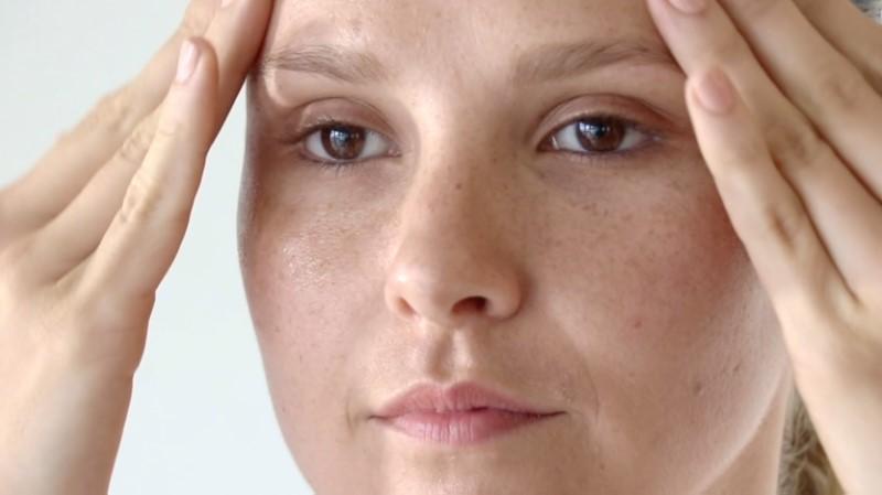 A close-up of a person gently massaging their forehead with both hands, applying Esse Hyaluronic Serum. The individual has light skin, brown eyes, and a calm expression.