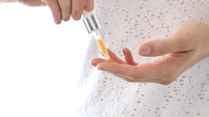 Close-up of a person dispensing Esse Repair Oil from a dropper onto their fingertip. The individual is adorned in a white lace top, and the softly lit background creates a clean and minimalist aesthetic.