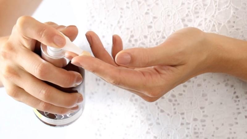 A person in a white lace top is applying the Esse Rich Body Moisturiser from a dark bottle with a white pump dispenser onto their fingers, poised as if about to rub the product onto their skin.