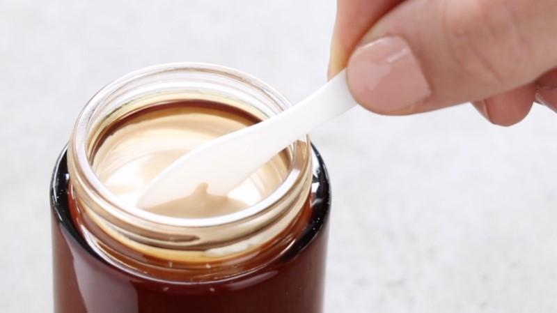 A hand with neatly manicured nails holds a white plastic spoon, dipping it into an open jar filled with the beige-colored creamy Esse Ultra Moisturiser. The jar appears to be made of dark glass. The background is a plain, light grey surface.
