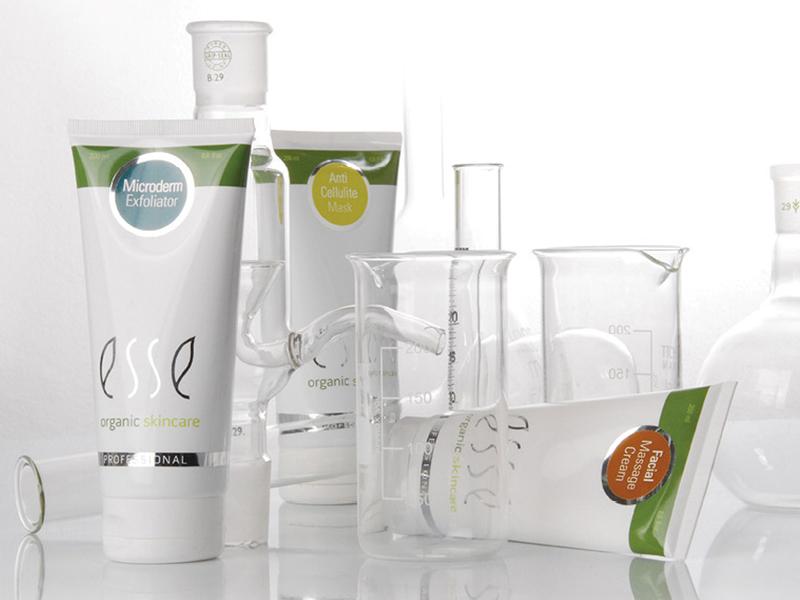 Various laboratory glassware and skincare product tubes from the brand "Esse" are arranged on a white surface. Visible products include a Microderm Exfoliator, Anti Cellulite Mask, and a Facial Moisturiser. The background is clean and minimalistic.