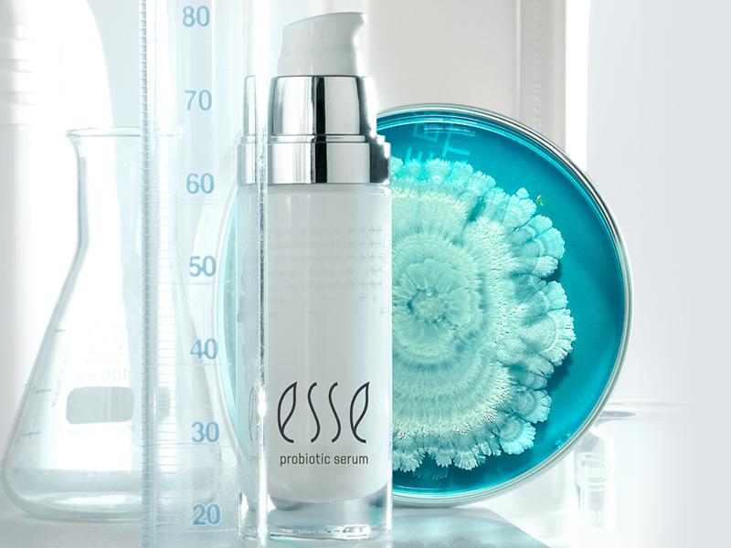 A white bottle of Esse probiotic serum with a silver pump sits in front of a blue petri dish with a white cultured microorganism inside. Lab glassware, including a graduated cylinder and a beaker, are in the background. The image has a clean, scientific ambiance.