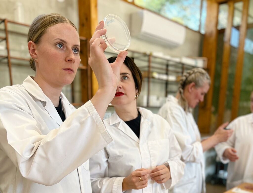 Four scientists wearing white lab coats work in a laboratory. Three are women; one holds and examines a petri dish, the second watches her, and the third conducts an experiment with equipment. The background shows lab shelves and a window.