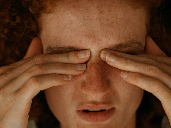 A person with freckles rubs their eyes with both hands, appearing to be tired or in discomfort. They have red curly hair and a close-up view focuses on their face and hands.