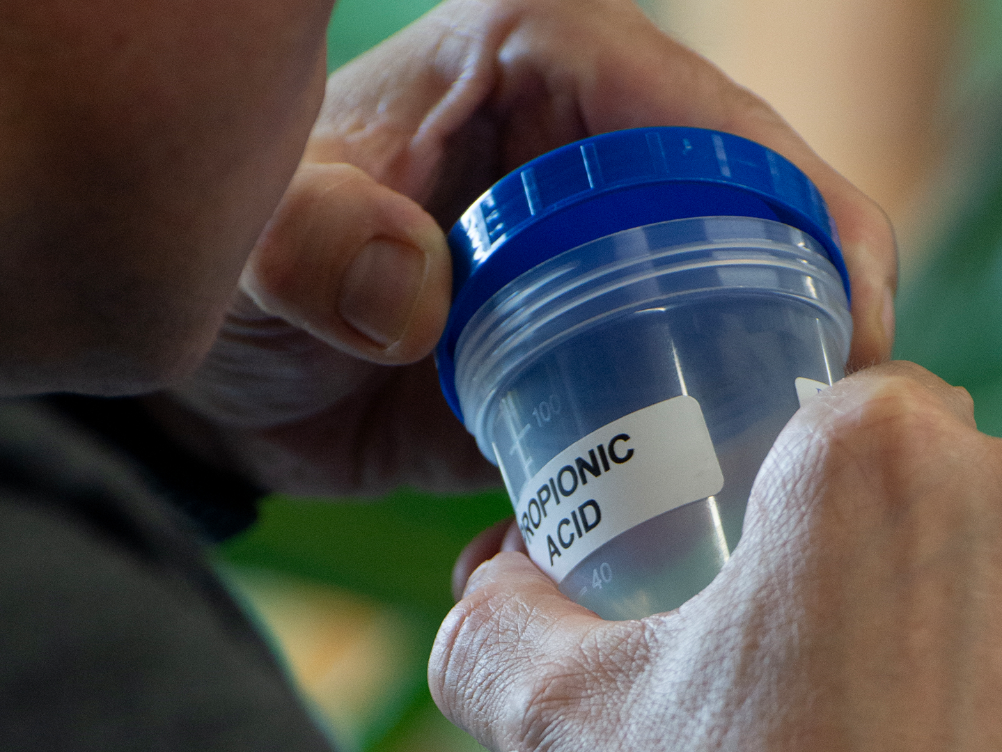 A person holds a transparent plastic container with a blue lid labeled "Propionic Acid." The focus is on the container and the person's hands, with a blurred background containing greenery.