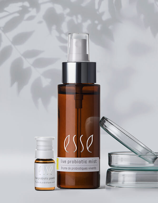 Two brown glass bottles of Esse Live Probiotic Mist sit on a white surface, one larger with a spray pump and one smaller with a white cap. Two glass petri dishes are beside them, with leafy shadows in the background.