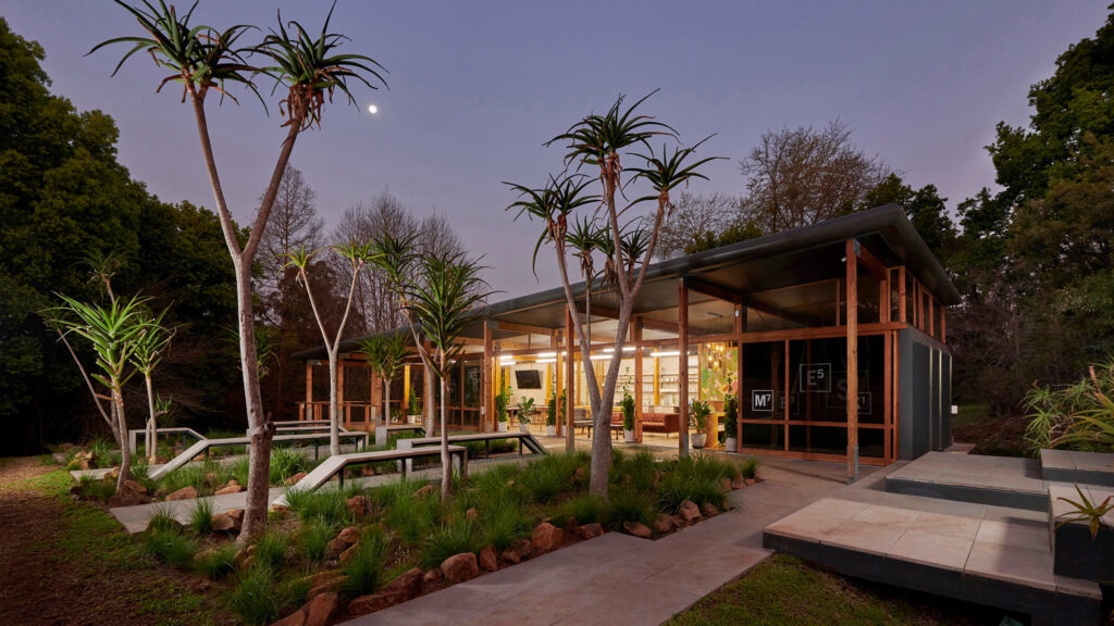A modern, glass-walled building is surrounded by trees and landscaping at dusk. Warm lights illuminate the interior, and a crescent moon is visible in the sky above. Pathways and desert plants are in the foreground.