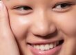 A close-up of a smiling person with clear, smooth skin, showing their teeth and part of their hand gently touching their face.